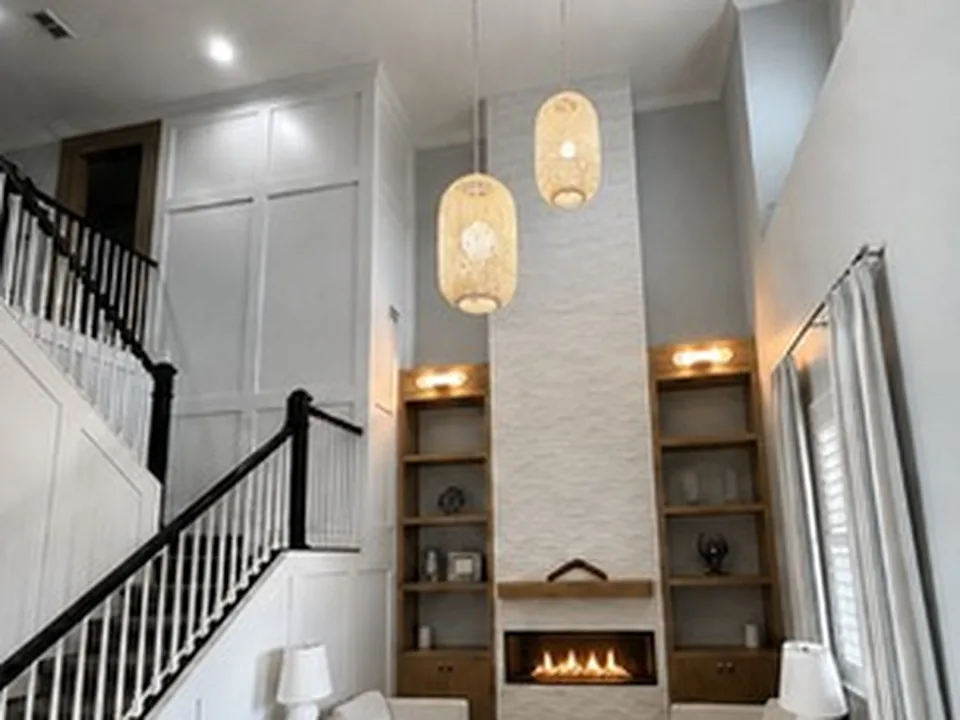 Two-story white stacked stone fireplace column with a linear fireplace and timber mantel, flanked by lit oak shelves beneath woven pendant lights